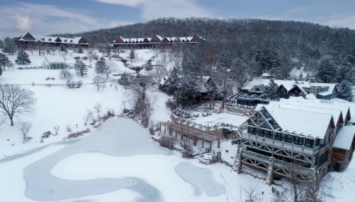 A snowy landscape featuring a frozen pond, rustic buildings, and a hillside with trees and cabins in the background.
