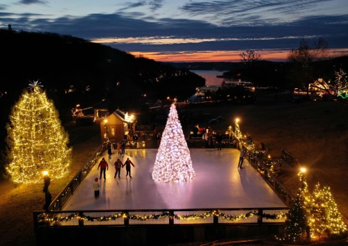 A festive ice skating rink with a large illuminated Christmas tree, surrounded by twinkling lights at sunset.