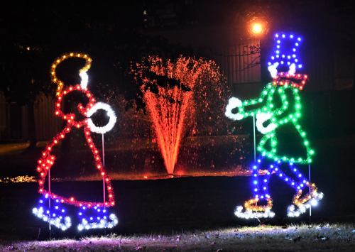 Colorful holiday lights depict a girl and a boy skating, with a fountain illuminated in the background.
