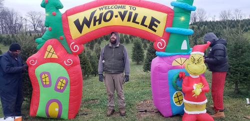 Inflatable "Welcome to Who-ville" arch with a Grinch figure, surrounded by people and Christmas trees in the background.