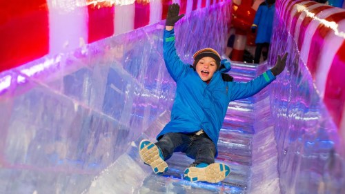 A joyful child in a blue coat slides down an ice slide, surrounded by colorful lights and ice sculptures.