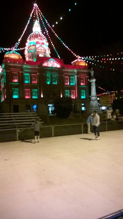 A festive ice skating rink at night, with a brightly lit building adorned in red and green lights in the background.