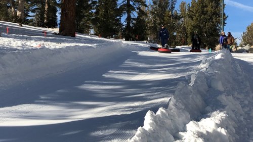 People tubing down a snowy slope surrounded by trees on a sunny day.