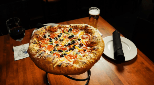 A large pizza topped with olives and peppers on a wooden table, accompanied by a glass of wine and a beer.
