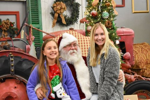 Two girls pose with Santa Claus in a festive setting, surrounded by holiday decorations and a vintage tractor.