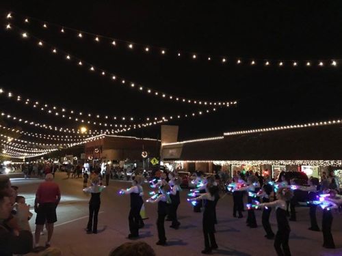 A nighttime scene of a festive parade with performers using glowing props under string lights. Crowds watch nearby.