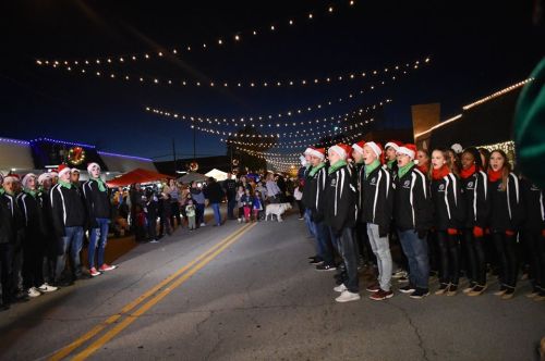 A group of carolers in Santa hats perform on a festive street decorated with lights during a holiday event.