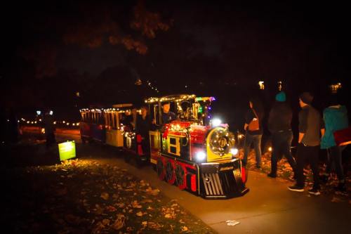 A festive train decorated with lights travels along a path, with people walking beside it at night.