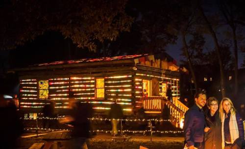 A cozy log cabin adorned with colorful lights at night, with three people smiling in the foreground.