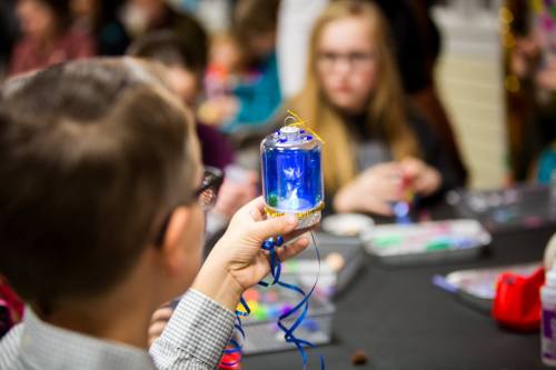 A child holds a glowing jar with colorful lights, surrounded by other kids engaged in a creative activity.