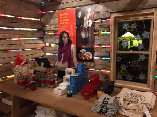 A woman stands behind a wooden table displaying various festive items in a cozy, decorated space.