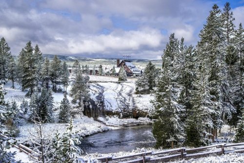 A snowy landscape with evergreen trees, a river, and a lodge in the background under a cloudy sky.