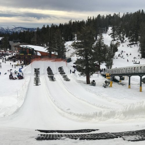 A snowy ski slope with people enjoying winter activities, surrounded by trees and a cloudy sky.