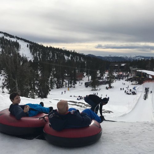 Two people sit on snow tubes at a winter resort, with snowy hills and trees in the background.