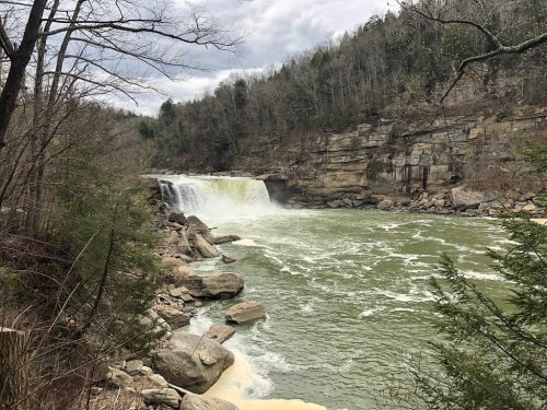 A waterfall cascades over rocky cliffs into a green river, surrounded by trees and a cloudy sky.