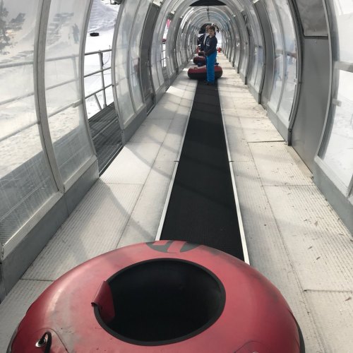 A snowy tubing area featuring a clear, curved tunnel and red inflatable tubes lined up on a conveyor belt.