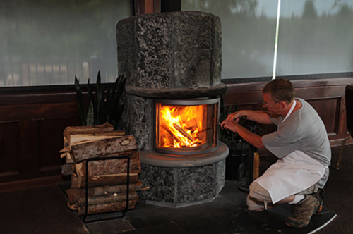 A person in an apron adds logs to a stone fireplace, with flames visible inside and firewood stacked nearby.