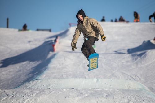 A snowboarder in a brown jacket jumps off a snow ramp against a clear blue sky. Snow-covered slopes are in the background.