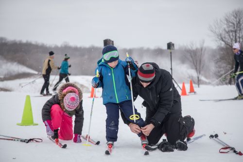 A parent helps a child with ski equipment while another child plays nearby in a snowy landscape with skiers in the background.