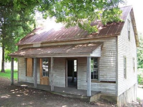 A weathered wooden house with a rusty metal roof, featuring boarded windows and surrounded by trees.