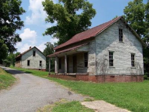 Two weathered, abandoned houses with peeling paint, surrounded by greenery and a gravel path under a blue sky.