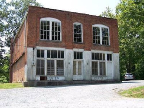 Abandoned brick building with large windows, surrounded by trees and gravel driveway.