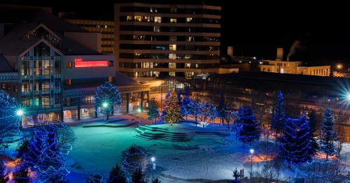 A winter scene at night featuring a park illuminated with blue lights and surrounding buildings in a city.
