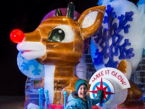 A child smiles next to a large ice sculpture of Rudolph the Red-Nosed Reindeer with a glowing button.