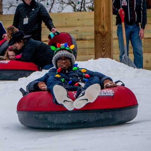 A child in a colorful hat rides a snow tube down a snowy slope, surrounded by other people enjoying winter activities.