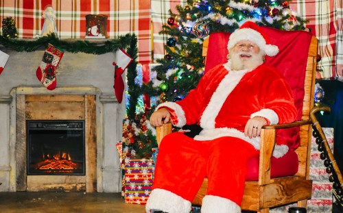 Santa Claus in a red suit sits in a cozy chair by a decorated Christmas tree and a fireplace, smiling warmly.