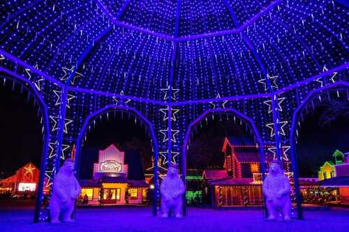 A festive display of colorful lights in a gazebo, featuring bear sculptures and decorated buildings in the background.