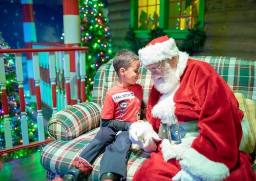 A child sits on a couch with Santa Claus, sharing a moment in a festive, decorated setting.