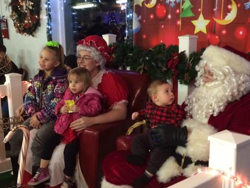 A festive scene with Santa, Mrs. Claus, and three children sitting together, surrounded by holiday decorations.