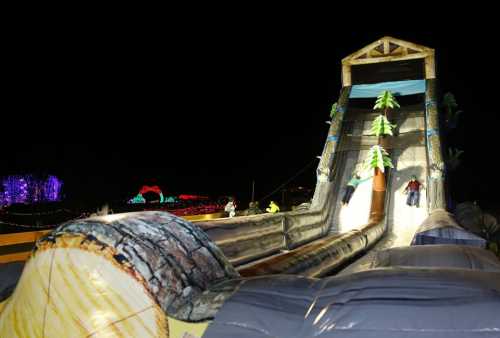 Two children slide down a large inflatable slide decorated like a log, surrounded by colorful lights at night.