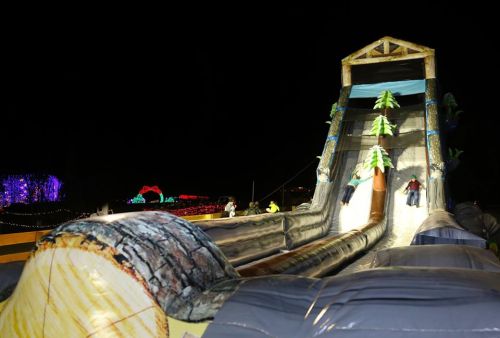Two children slide down a large inflatable slide decorated like a log, surrounded by colorful lights at night.