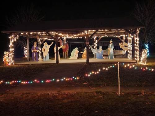 A lit pavilion displays a nativity scene with figures and colorful lights, set against a dark night sky.