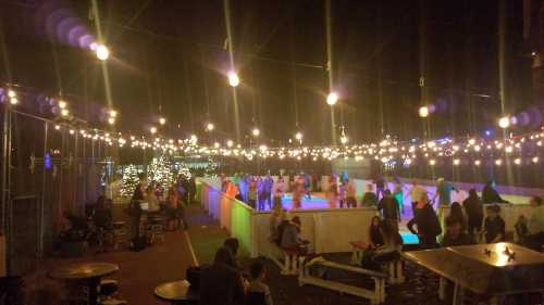 A lively outdoor ice skating rink at night, illuminated by string lights, with people skating and socializing.