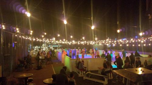 A lively outdoor ice skating rink at night, illuminated by string lights, with people skating and socializing.