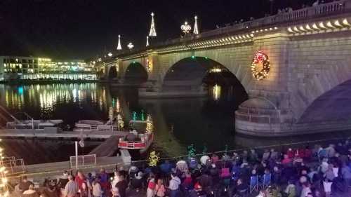 A festive bridge lit up at night, with a crowd gathered by the water and a boat passing underneath.