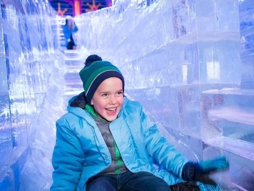 A joyful child in a blue coat and hat slides down an icy tunnel, surrounded by shimmering ice blocks.