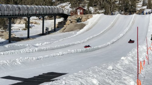 Two snow tubing lanes with people sliding down, surrounded by snow and trees, with a ski lift in the background.