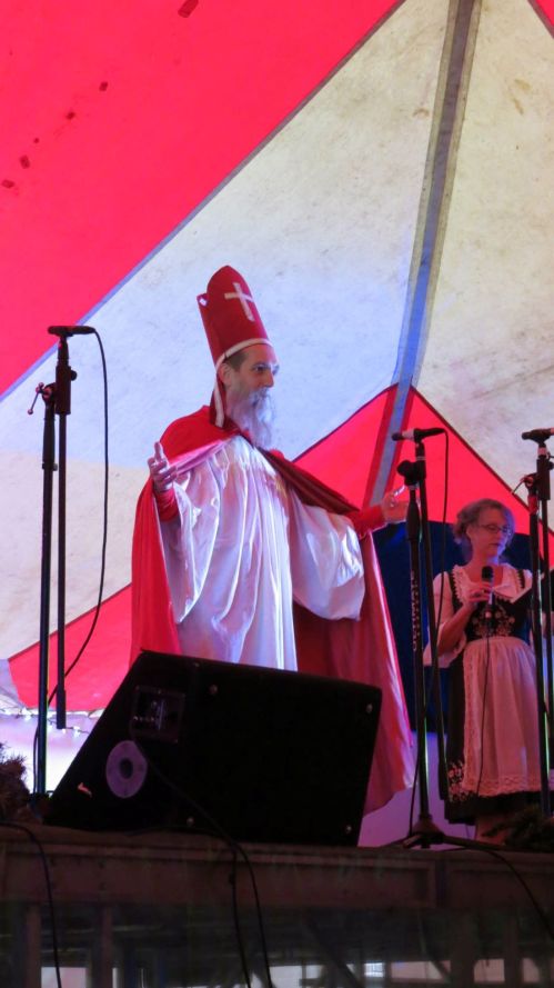A man in a red and white robe and hat stands on stage, gesturing, with a woman in traditional attire beside him.