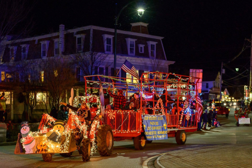 A festive parade float decorated with lights, featuring people in holiday attire and a snowman, moving through a town at night.