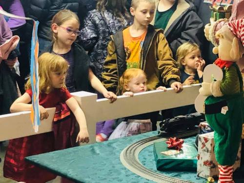 A group of children watches a festive display with a train set and decorations, eagerly leaning forward.