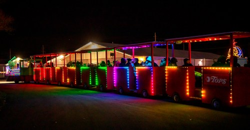 A brightly lit train with colorful lights, carrying passengers at night, with a festive atmosphere in the background.