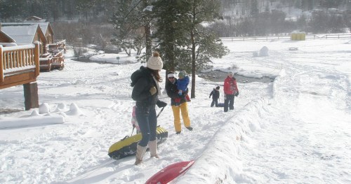 A group of people walking in the snow, carrying sleds near wooden cabins and trees in a winter landscape.