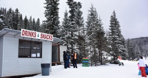 A snowy scene featuring a small snack shack with a "Drinks & Snacks" sign, surrounded by evergreen trees.