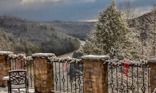 Snow-covered landscape with a river, trees, and a stone railing adorned with festive decorations.