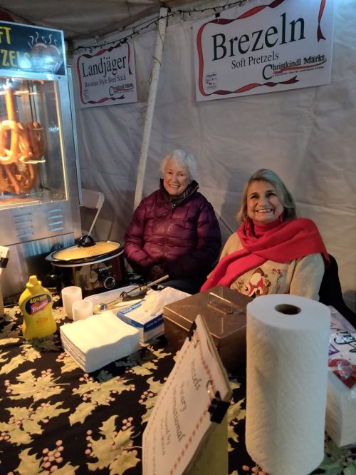 Two women smile behind a booth selling soft pretzels, with mustard and snacks on the table, in a festive setting.