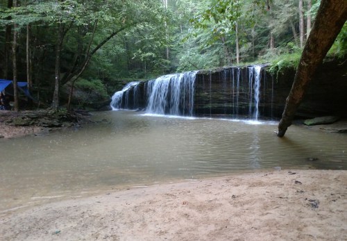 A serene waterfall cascades into a calm pool, surrounded by lush greenery and sandy banks.